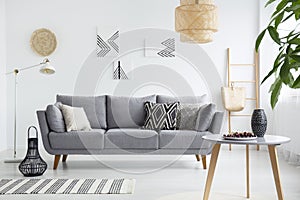 Real photo of a scandi living room interior with cushions on gray couch, cherries on wooden table and bag on a ladder