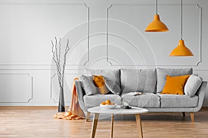 Real photo of simple living room interior with orange lamps, pillows and grey sofa