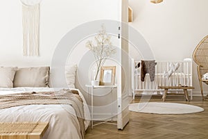 White bedroom interior with bedside table with plant, poster and mug and open door to baby room with rug and white c photo