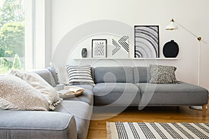 Real photo of white Scandi sitting room interior with metal lamp, corner sofa with cushions and modern art posters