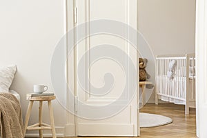 Real photo of wooden stool with book and tea mug standing next to white door to kid room interior with crib