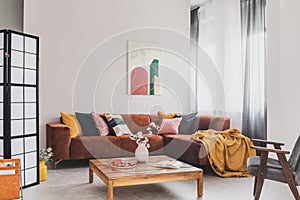 Real photo of a yellow blanket lying on a brown corner sofa in bright living room interior with a wooden table and a gray chair