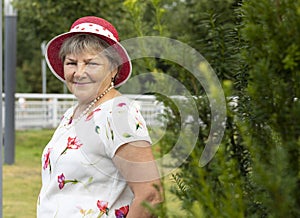 Real Smiling Senior Woman In Hat In Sunny Day Looks At Camera. Green Trees, Park on