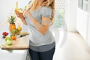 Real women have curves. Curvy young woman eating a banana in her kitchen at home.