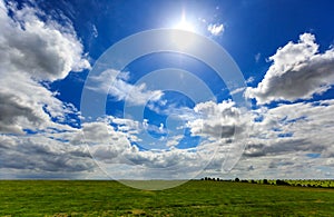 Realistic blue cloudy sky with bright sun over green meadow field