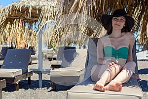 Realistic portrait of a young woman in a green swimsuit and hat, sitting on a chaise longue under a straw umbrella