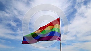 Realistic rainbow flag of an LGBT organization waving against a blue sky, clouds . LGBT pride flags include lesbians