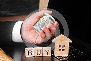 The realtor`s hand holds a bundle of money behind the miniature house. The concept of buying and selling real estate, investment.