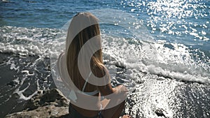 Rear back view of unrecognizable young girl in bikini sitting on stone near seashore and looking into distance on sunny