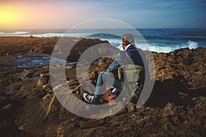 Rear portrait of a relaxed male traveler sitting on the rocky cliff and enjoying the view of Atlantic ocean at sunset