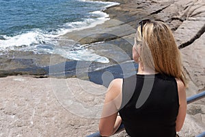 Rear profile view of an attractive Caucasian woman in a black top leaning against a metal rail and admiring the frothy waves