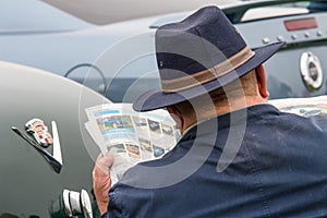 Rear shot of a caucasian man in a hat sat beside classic cars reading adverts for classic cars in a newspaper.  Over shoulder shot