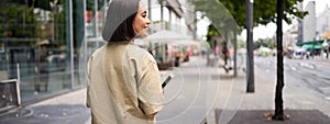 Rear shot of young woman walking in city, going down the street and smiling, holding smartphone. View from behind