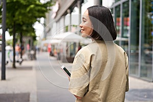 Rear shot of young woman walking in city, going down the street and smiling, holding smartphone. View from behind