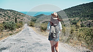 Rear view active backpacker woman in hat walking on path to sea surrounded by mountains