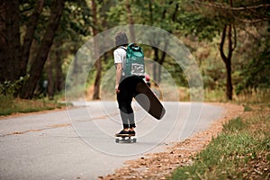 Rear view of active young woman riding skateboard with wakeboard in her hand and backpack on her shoulders.