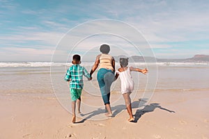 Rear view of african american grandmother holding boy and girl's hands running at beach against sky