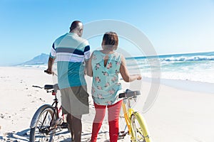Rear view of african american retired senior couple with bicycles at beach on sunny day