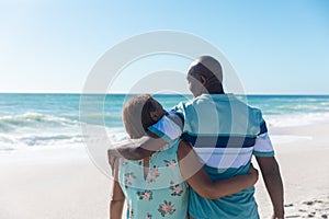 Rear view of african american senior couple standing with arms around at beach with copy space