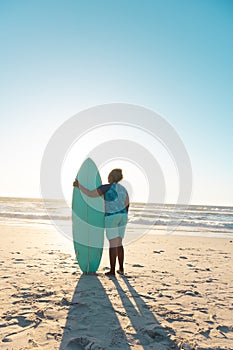 Rear view of african american senior woman with surfboard looking at sea under blue sky at sunset