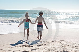 Rear view of african american siblings holding hands while running towards sea at beach on sunny day