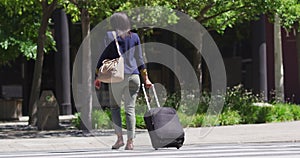 Rear view of african american woman wearing face mask crossing road wheeling suitcase