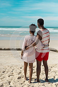 Rear view of african american young couple with arms around looking at sea while standing on beach