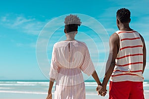 Rear view of african american young couple holding hands and looking at sea while standing on beach