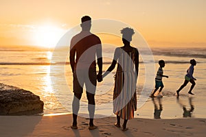 Rear view of african american young couple looking at playful children running on beach against sky