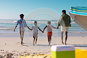 Rear view of african american young parents holding children's hands running at beach against sky