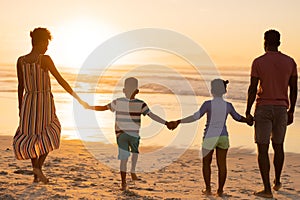 Rear view of african american young parents holding daughter and son\'s hands while walking on beach