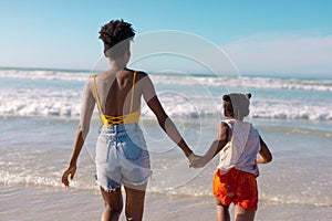 Rear view of african american young woman holding daughter's hands and running in sea against sky