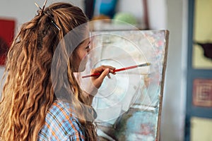 Rear view of an artist female standing next to the easel with canvas painting something in her art studio. A student young woman