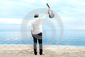 Rear view of asian businessman in hat and white clothes holding guitar on the beach