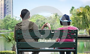 Rear view of Asian couples holding hands and sitting apart on green garden bench in public park area