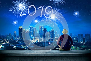 Rear view of asian traveler man sitting on the rooftop looking at 2019 from sparkling fireworks on the city at night