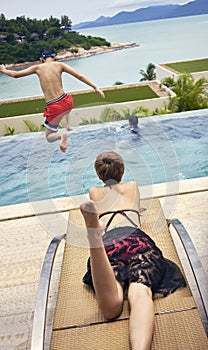 Rear view of Asian woman lying on stomach sunbathing on deck chair while kid jumping into pool