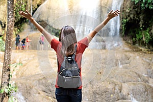 Rear view of Asian woman standing in front of waterfall with her hands raised.