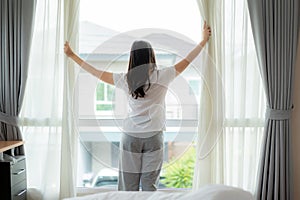 Rear view of Asian woman waking up in her bed fully rested opening window curtains and looking through the window in bedroom at