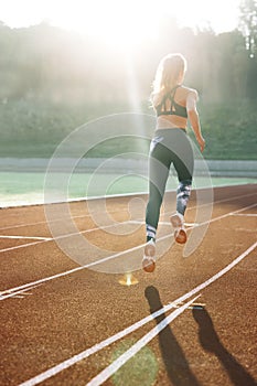 Rear view of athletic woman running on track stadium at summer morning light. Fit woman running on racetrack during