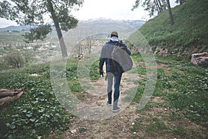 Rear view of a backpacked male tourist walking on the forest foot path in early spring nature, discovering mountains