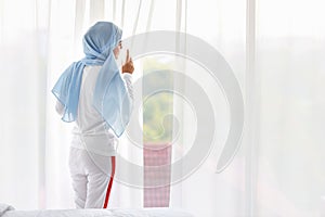Rear view beautiful asian Arab woman wearing white sleepwear looking away from window after getting up in the morning at sunrise.