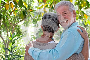 Rear view of a beautiful senior couple walking in the woods tenderly embraced while the man looks at the camera. Smiling elderly