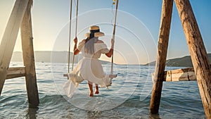 Rear view of beautiful woman in long dress having fun swinging on wooden swing on the sea shore. Tourism, summertime