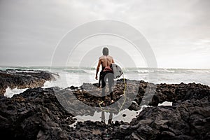 Rear view brunette boy standing in the black swimsuit with naked torso with a white surf in his hands on the shore