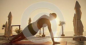 Rear view of a brunette girl in a black sports summer uniform doing plank exercises on a red mat on the Sunny seashore