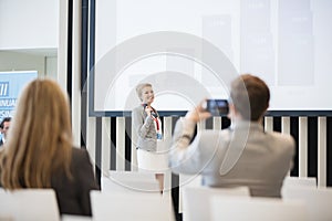 Rear view of businessman photographing female public speaker in seminar hall