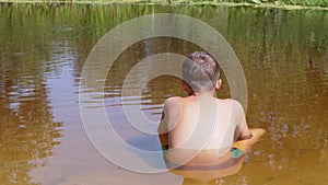 Rear View of a Child sitting on Sand Up to Waist in a Transparent, Shallow River