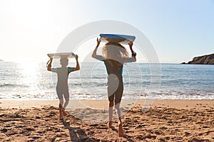 Rear View Of Children In Wetsuits Carrying Bodyboards On Summer Beach Vacation Having Fun By Sea