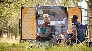 Rear View Of Couple Camping In Countryside With RV Drinking Coffee By Outdoor Fire With Pet Dog